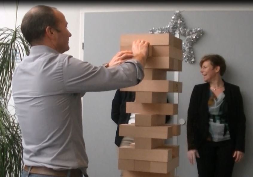 Een man bouwt een gigantische Jenga-toren, terwijl iemand anders binnen met een glimlach toekijkt.
