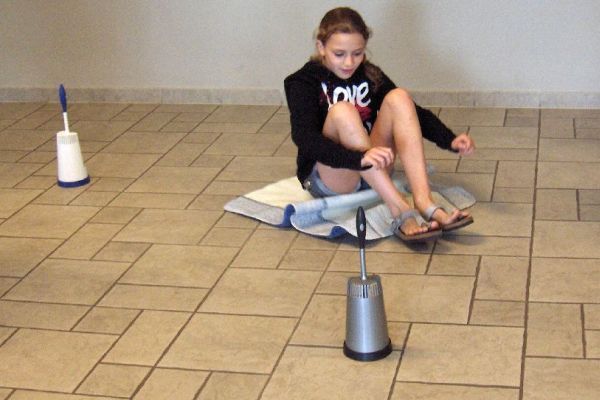 Een meisje zit op een handdoek en strikt haar sandalen. Twee toiletborstels staan ​​vlakbij op de tegelvloer.