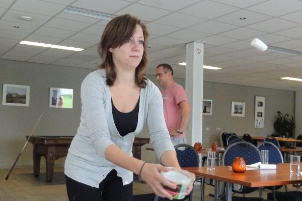 Een vrouw gooit een wit voorwerp, blijkbaar tijdens een spelletje binnenshuis. Een man kijkt toe op de achtergrond; tafels en stoelen zijn zichtbaar.
