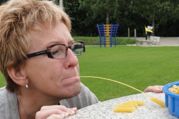 Vrouw met bril probeert een sliert spaghetti op te eten, vlakbij een kom pasta in de buitenlucht.