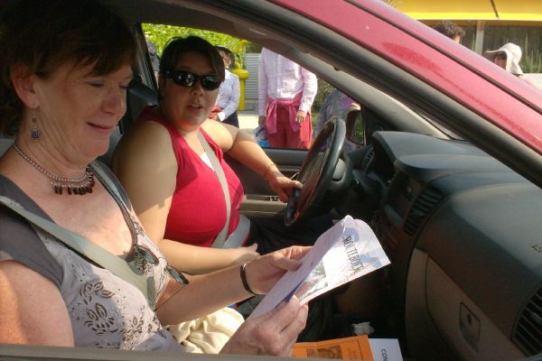 Twee vrouwen in een rode auto, de ene bestuurt de auto, de andere leest de krant. Buiten zijn andere mensen en een geel gebouw zichtbaar.