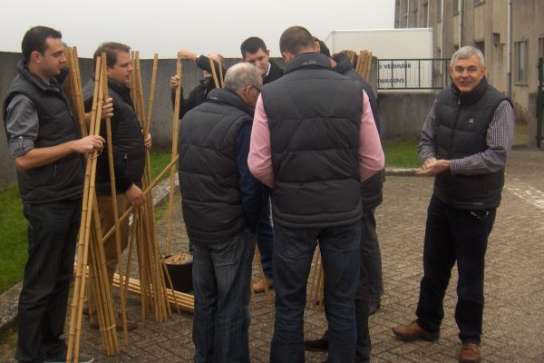 Een groep mannen in hesjes staat buiten, houten palen vast te houden en te onderzoeken, vlakbij een gebouw.