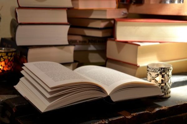 Een open boek ligt op een tafel, met stapels boeken op de achtergrond. Een kaars staat ernaast.