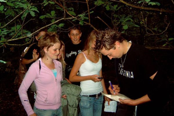Een groep mensen in een donker bos. Een man ondertekent iets, terwijl verschillende anderen toekijken.