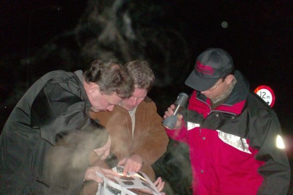 Drie mannen staan ​​dicht bij elkaar in het donker, mogelijk kijkend naar iets waar stoom vanaf komt; een van hen houdt een zaklamp vast en draagt ​​een rode jas.
