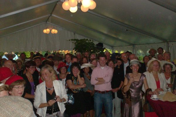 Een grote groep mensen onder een witte tent, mogelijk bij een evenement, velen dragen hoeden.