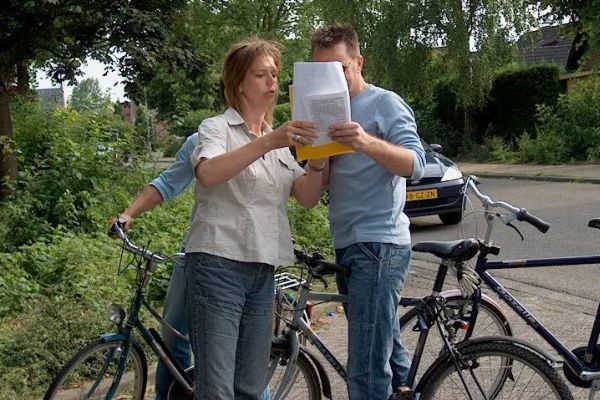 Een vrouw en een man bekijken buiten papieren; op de achtergrond zijn fietsen, een auto en groen te zien.