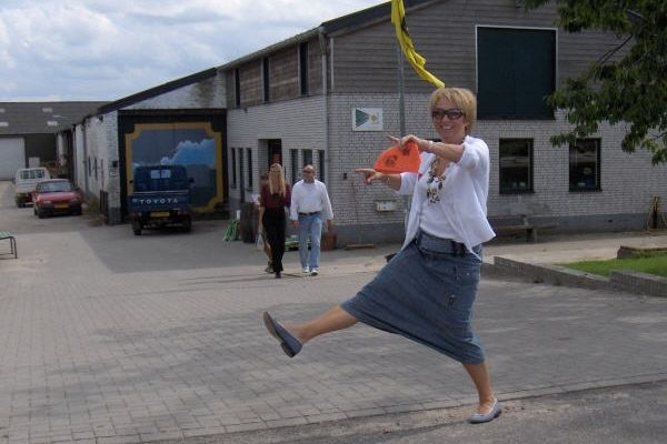 Een vrouw trapt buiten voor een gebouw tegen een oranje voorwerp; op de achtergrond lopen twee mensen.