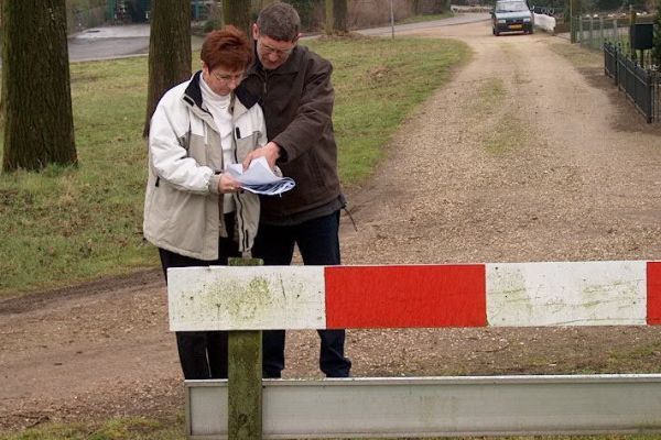 Een man en een vrouw lezen kranten bij een rood-witte afscheiding op een grindweg, met een voertuig in de verte.