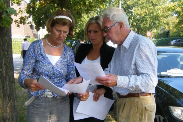 Drie mensen bekijken documenten in de buitenlucht. Eén draagt ​​een blauw vest, de middelste een zwarte blazer en de laatste een lichtblauw overhemd.
