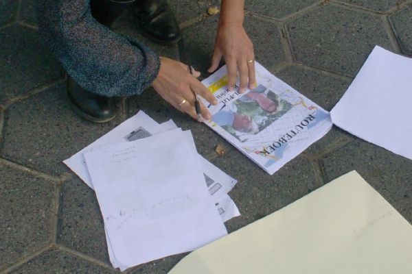 Een persoon vouwt een krant op een met bakstenen bestrate ondergrond, omringd door blanco witte en gebroken witte vellen papier en plastic zakken.
