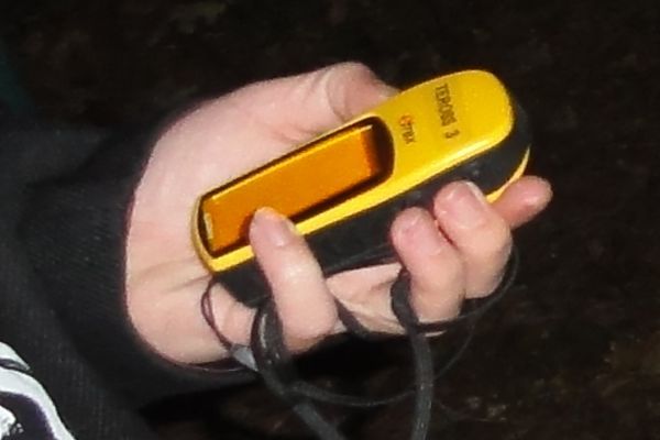 De hand van een persoon houdt een geel GPS-apparaat vast met een oranje scherm en een zwarte band.