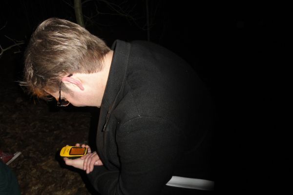 Persoon in donkere jas kijkt naar een mobiel apparaat, buiten in de nacht.