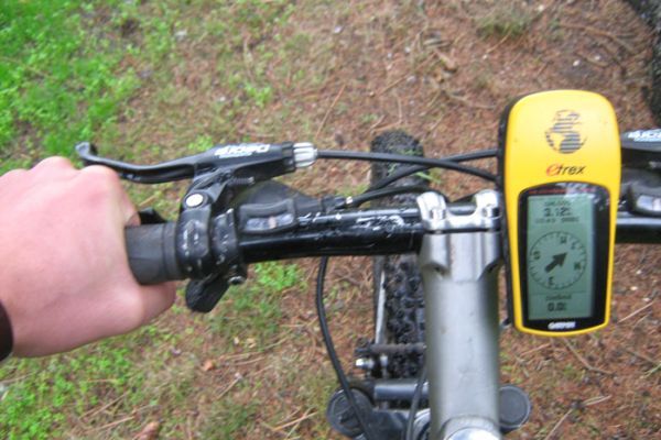 Hand die een fietsstuur vasthoudt met daarop een geel GPS-apparaat, op een fietspad.