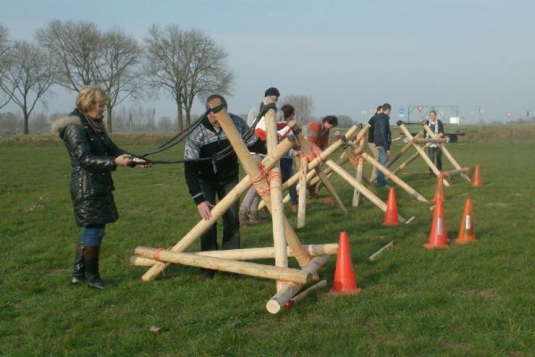 Mensen bedienen zelfgemaakte houten katapulten op een grasveld; oranje kegels markeren de doelen.