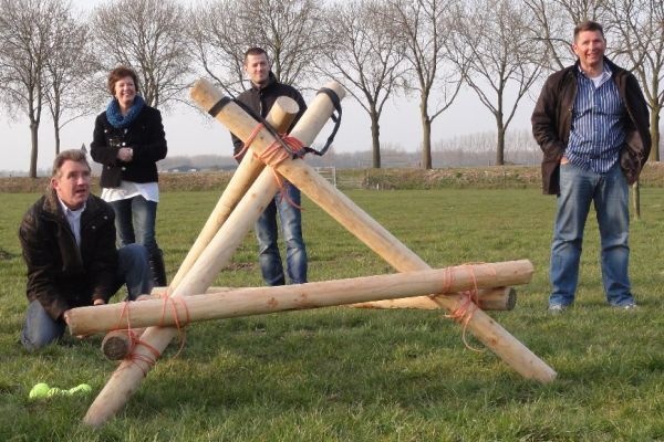 Vier mensen met een houten constructie in een veld. Eén knielt, de anderen staan.