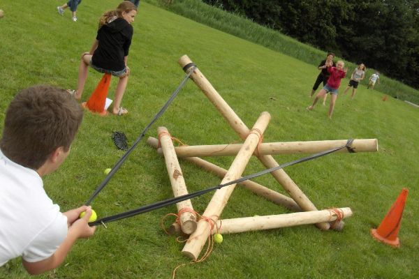 Een jongen richt een grote katapult op een doelwit. Andere mensen staan ​​op een grasveld.