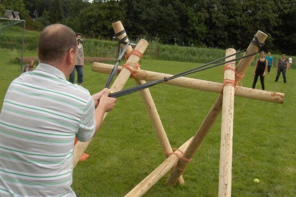 Een man richt een grote katapult op een doelwit in een grasveld; anderen kijken toe.