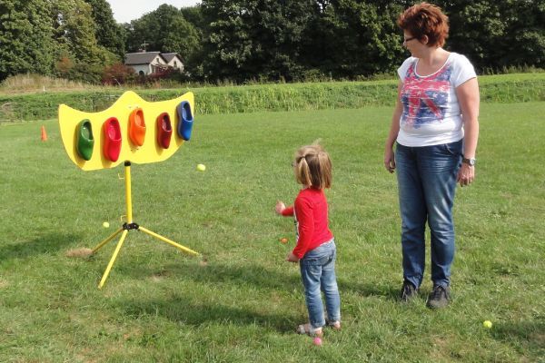 Een meisje gooit een bal tijdens een kleurrijk kermisspel; een volwassen vrouw kijkt toe vanaf een grasveld.