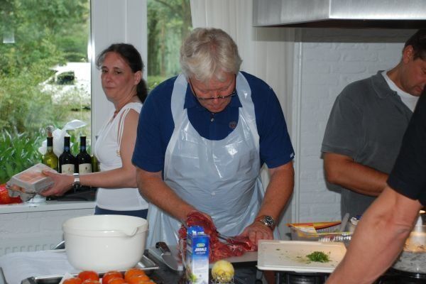 Een man met een schort snijdt vlees in de keuken; een vrouw en een andere man staan ​​er vlakbij.