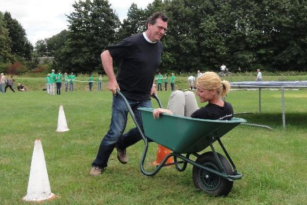 Een man duwt een vrouw in een kruiwagen door een grasveld met kegels. Anderen kijken op de achtergrond toe.