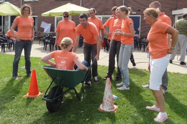 Mensen in oranje shirts spelen een spel in de buitenlucht. Eén persoon zit in een kruiwagen die door anderen wordt voortgeduwd, er staan ​​oranje kegels in het gras.