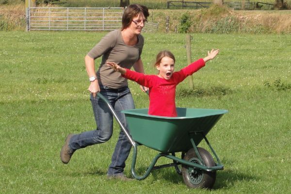 Een vrouw duwt een kind in een groene kruiwagen over een grasveld. Het kind heeft zijn armen enthousiast omhoog.