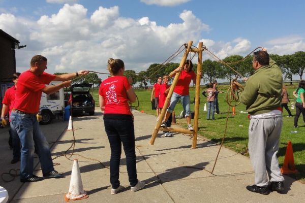 Mensen spelen een teambuildingspel in de buitenlucht. Eén persoon balanceert op een houten constructie, terwijl anderen touwen vasthouden.