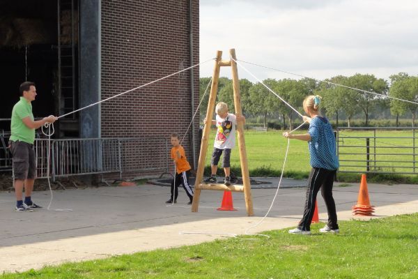 Kinderen spelen een touwloopspel, onder begeleiding van twee volwassenen. Buiten, vlakbij een bakstenen gebouw en gras.