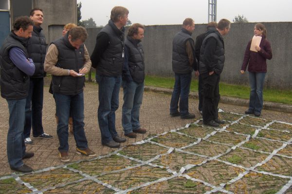 Een groep mensen bekijkt een raster op de grond in de buitenlucht. Sommigen kijken naar beneden, één persoon houdt aantekeningen vast.