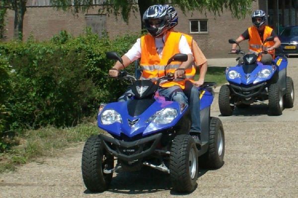 Twee personen, gekleed in helmen en hesjes, rijden op blauwe quads over een verhard pad.