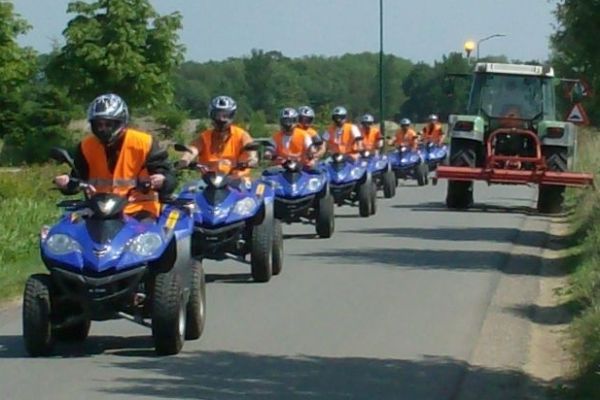 Een rij mensen op blauwe quads volgt een tractor op een geasfalteerde weg; ze dragen allemaal oranje hesjes en helmen.