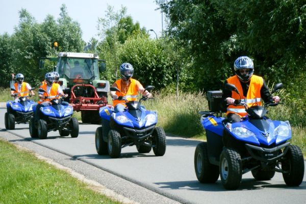 Vier mensen op blauwe quads, met helmen en oranje hesjes aan, rijden achter een tractor over een weg.