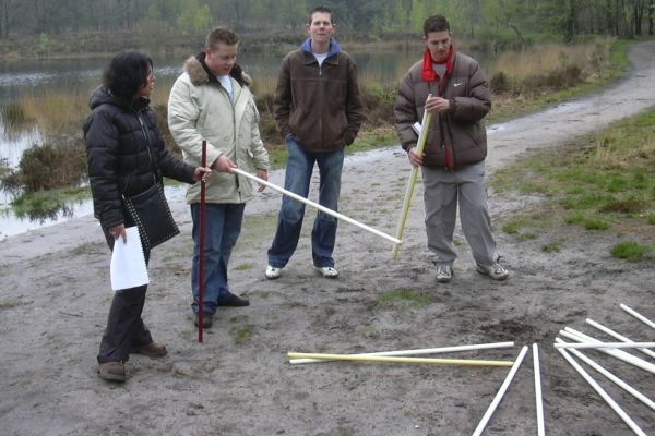 Vier mensen buiten met stokken; een vrouw wijst, drie mannen kijken toe, modderige grond.