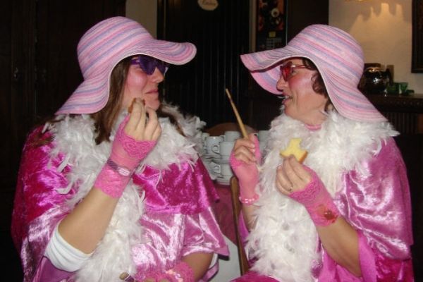 Twee mensen in roze kostuums met grote hoeden, boa's en brillen, die binnenshuis met elkaar in gesprek zijn.
