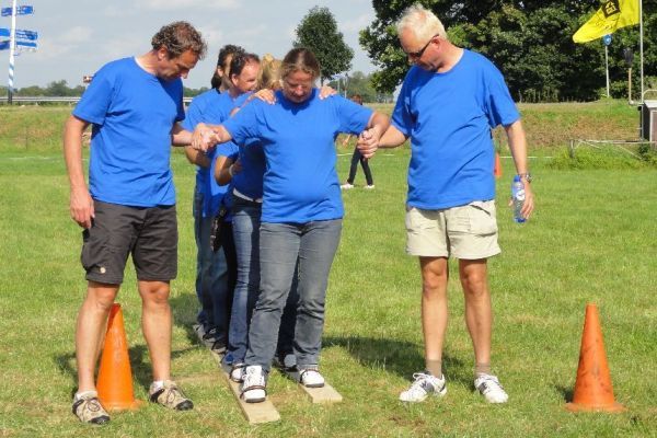 Mensen in blauwe shirts doen mee aan een teambuildingsoefening in de buitenlucht, lopend over houten planken.