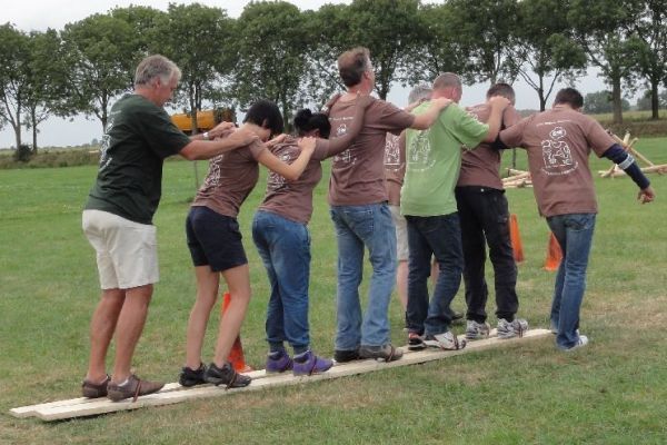 Een groep mensen, arm in arm, loopt buiten over een houten plank als teambuildingsoefening.