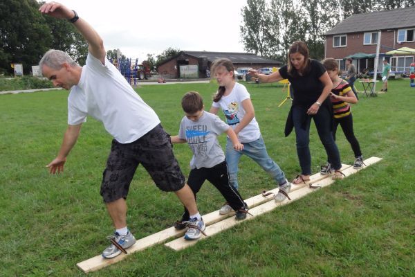 Mensen lopen over houten planken over een grasveld, waarschijnlijk spelend een spel, waarbij één persoon zijn armen uitstrekt en zo zijn evenwicht bewaart.