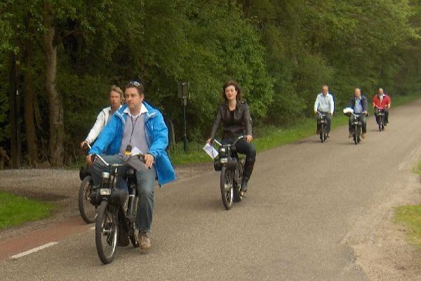 Een groep mensen rijdt op bromfietsen over een weg met bomen.