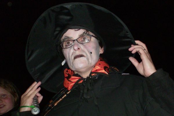 Persoon in een heksenpak met witte gezichtsmake-up, een zwarte hoed en een oranje sjaal.