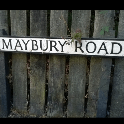 White rectangular road sign with black lettering against a greyish-brown wooden fence, depicting Maybury Road, Woking, Surrey, England, home of writer HG Wells 1895-1896.