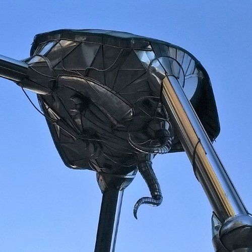 Coloured photo of the famous Martian tripod sculpture in Woking town centre, Surrey, England, taken from directly below the black metallic tripod 'hood' against a bright blue, cloudless sky.