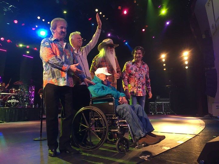Group of musicians on stage, some wearing colorful shirts, one in a wheelchair, waving to audience.