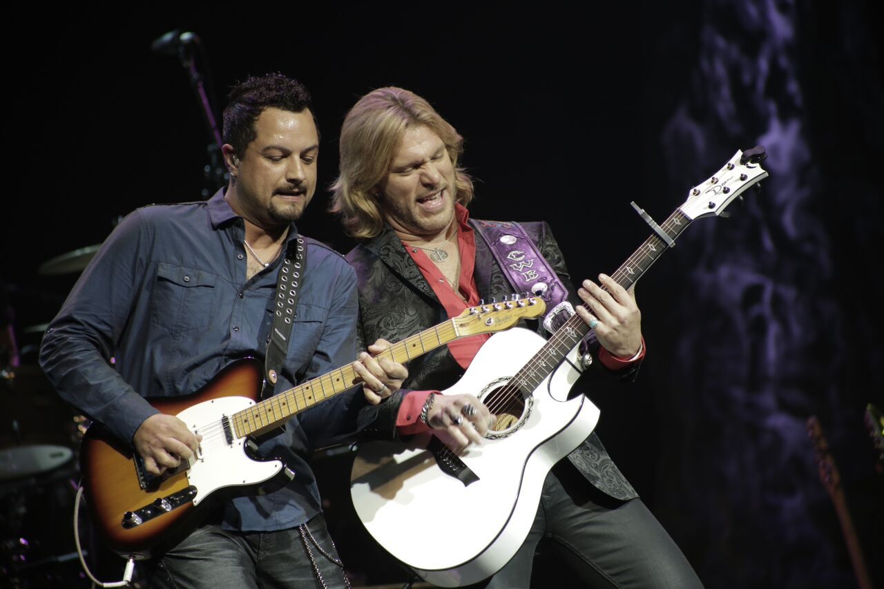 Two men playing guitars on stage, one with electric, one with acoustic, both singing.