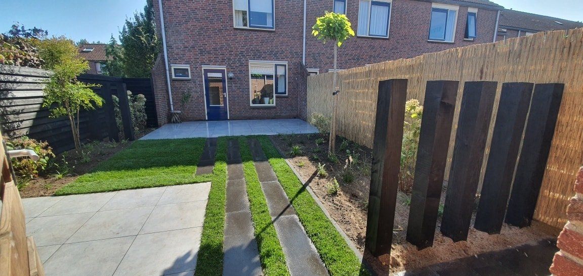 Achtertuin met betonnen pad, gras en bakstenen huis. Houten schutting en bomen op een zonnige locatie.