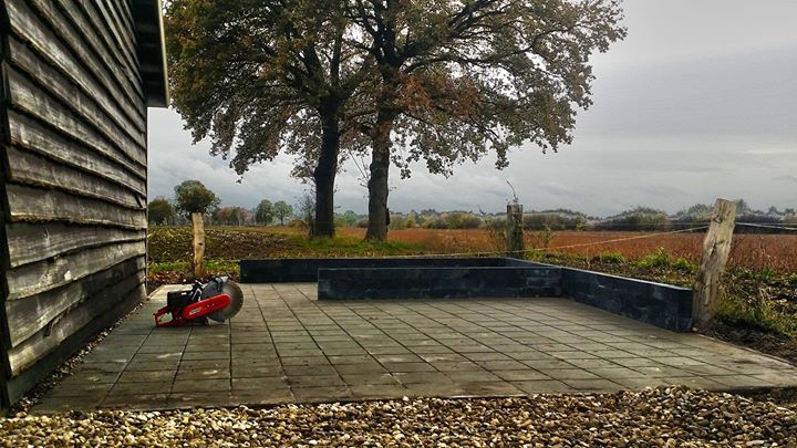 Buitenzicht van een patio met een rode grasmaaier, een verweerd gebouw, bomen en een veld onder een bewolkte hemel.