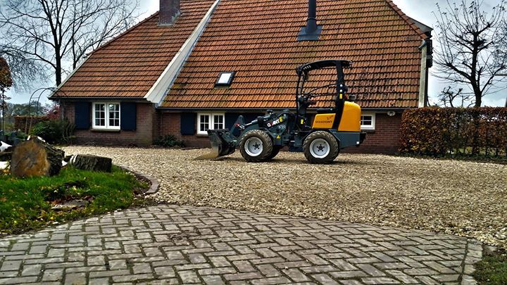 Een kleine geel-grijze shovel op een grindpad voor een bakstenen huis met een oranje dak.