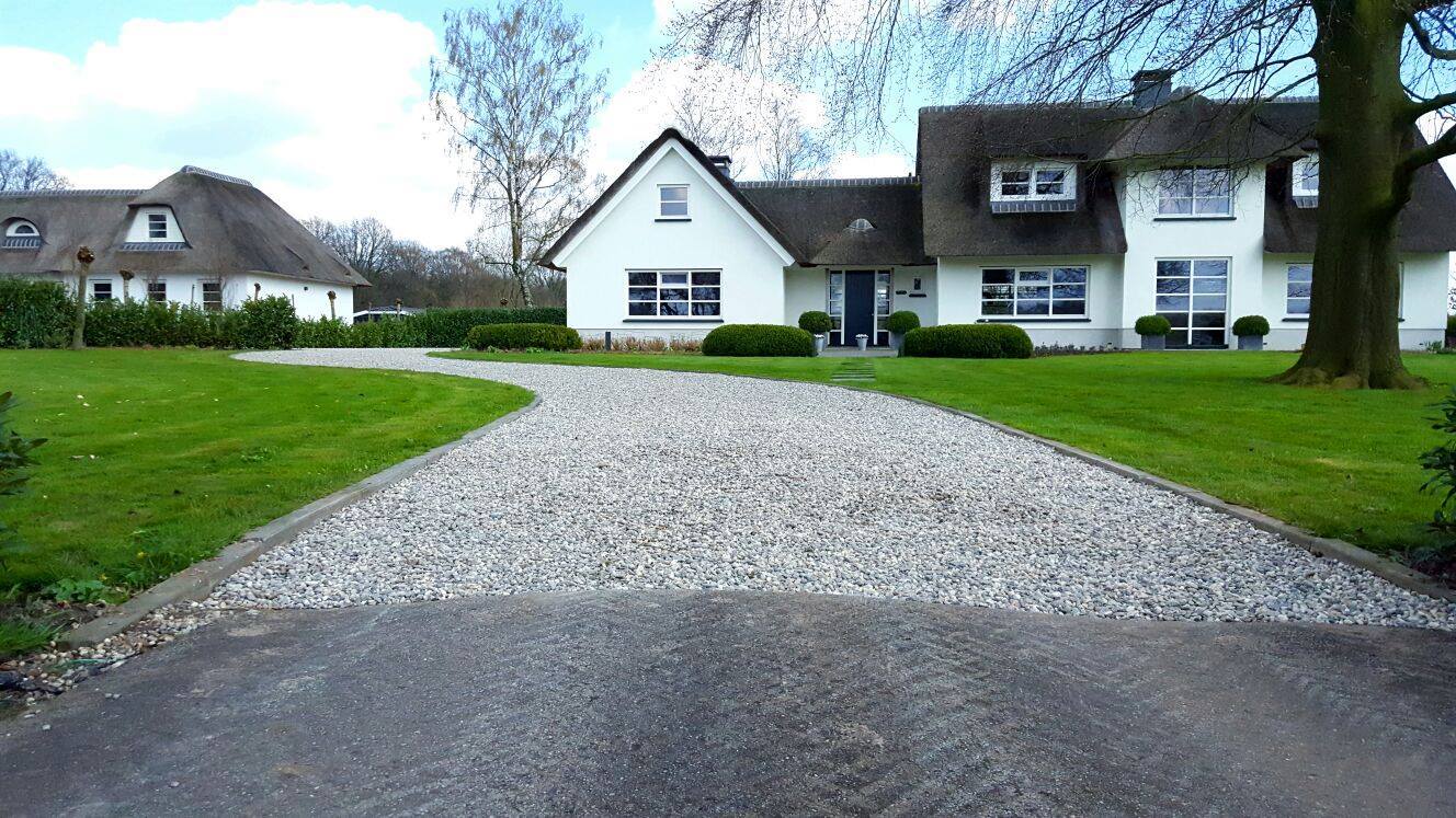Een grindpad leidt naar een wit huis met een rieten dak, omgeven door groen gras en bomen.