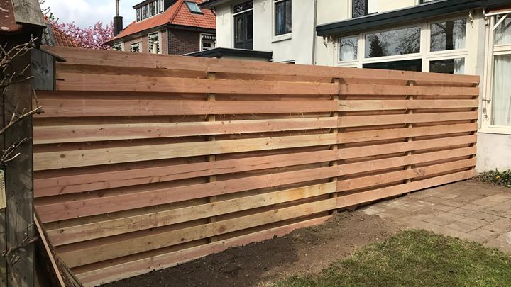 Een houten schutting van horizontale latten in een tuin met een gebouw op de achtergrond.