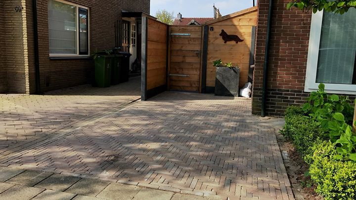 Een bakstenen oprit leidt naar een houten poort, geflankeerd door een bakstenen gebouw, een groene afvalcontainer en groen.